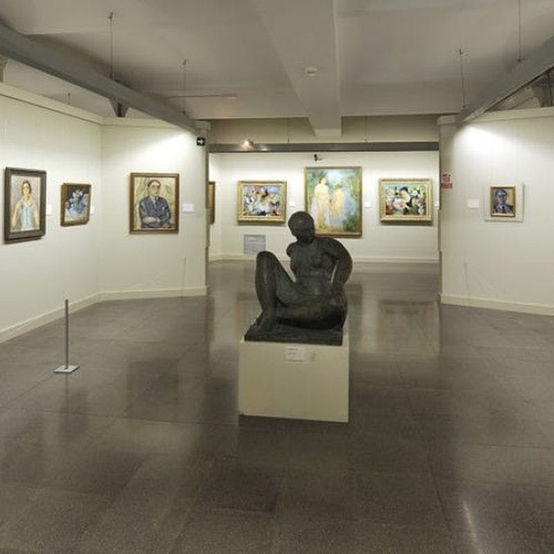 Intérieur d'une galerie d'art avec divers tableaux encadrés sur les murs et une sculpture de personnage assis sur un piédestal au centre.
