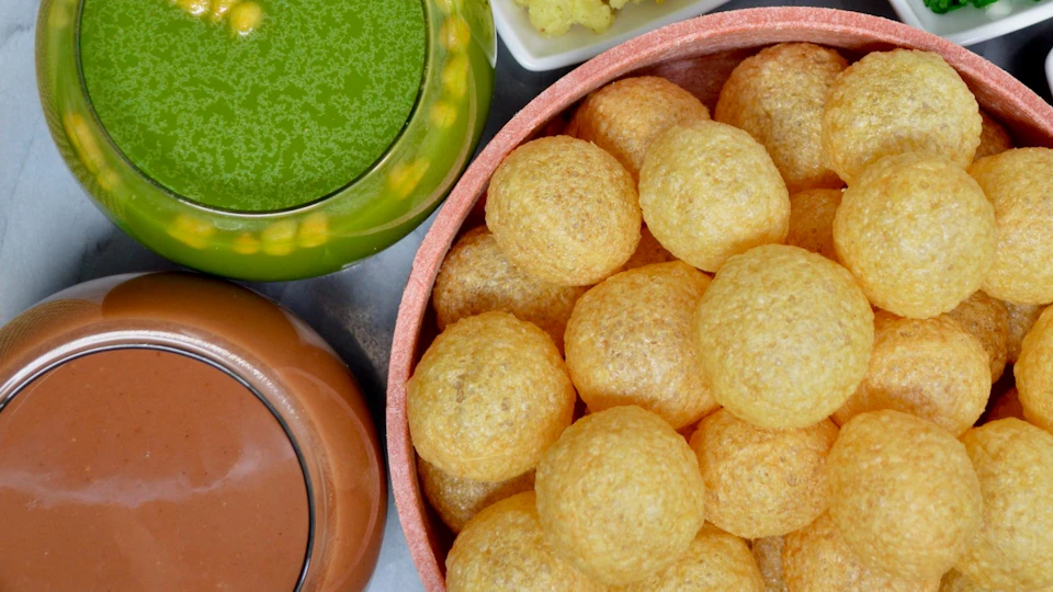 A basket of round, crispy puris beside containers of green chutney, brown sauce, and a small dish of yellow potatoes.