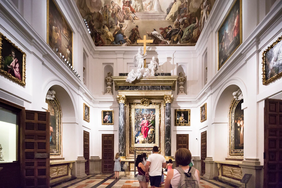 People viewing ornate paintings and sculptures in a grand gallery with a decorated ceiling and arched doorways.
