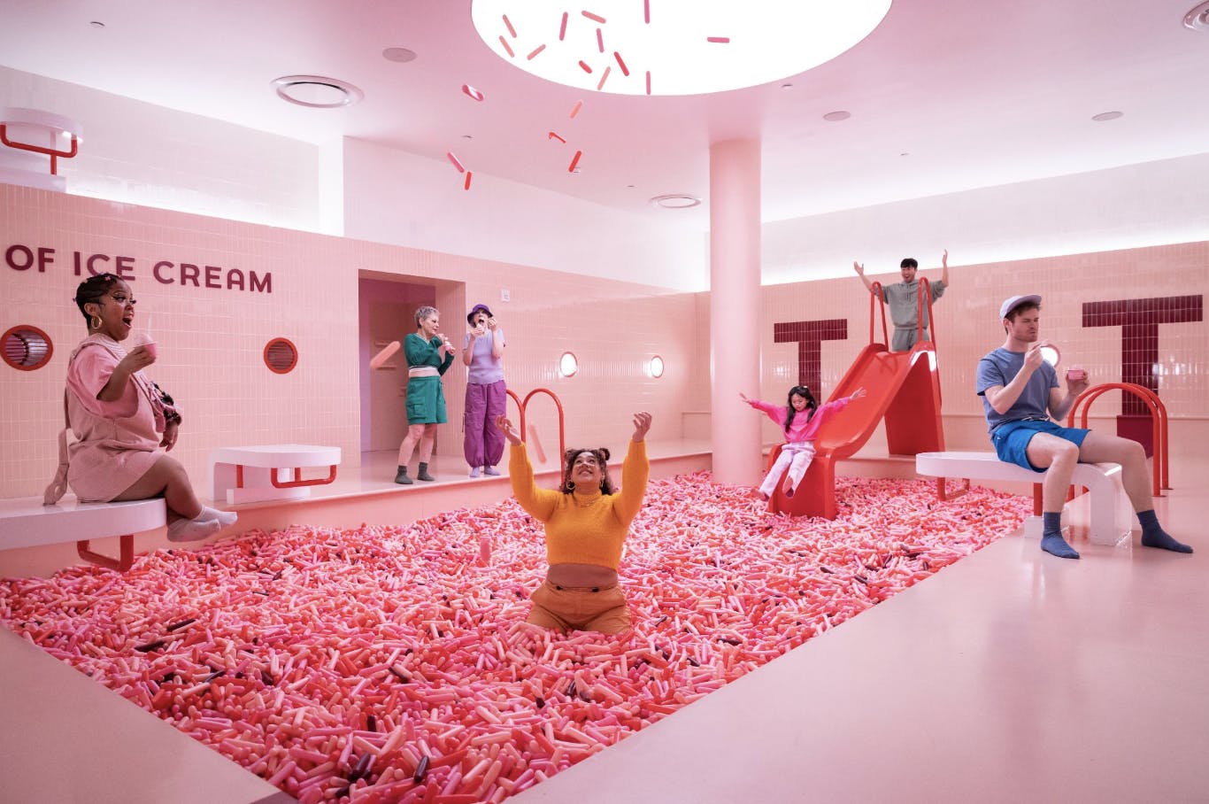 분홍색과 빨간색 볼 구덩이와 미끄럼틀이 있는 밝은 색상의 놀이방을 즐기는 사람들. 인공 소시지는 위에서 비가 내립니다.