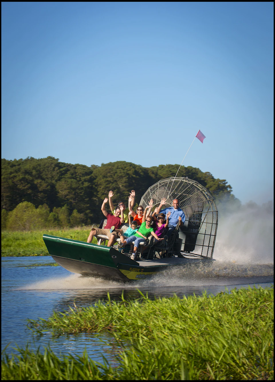A group of people on an airboat ride through a grassy waterway, raising their hands in excitement against a backdrop of trees.