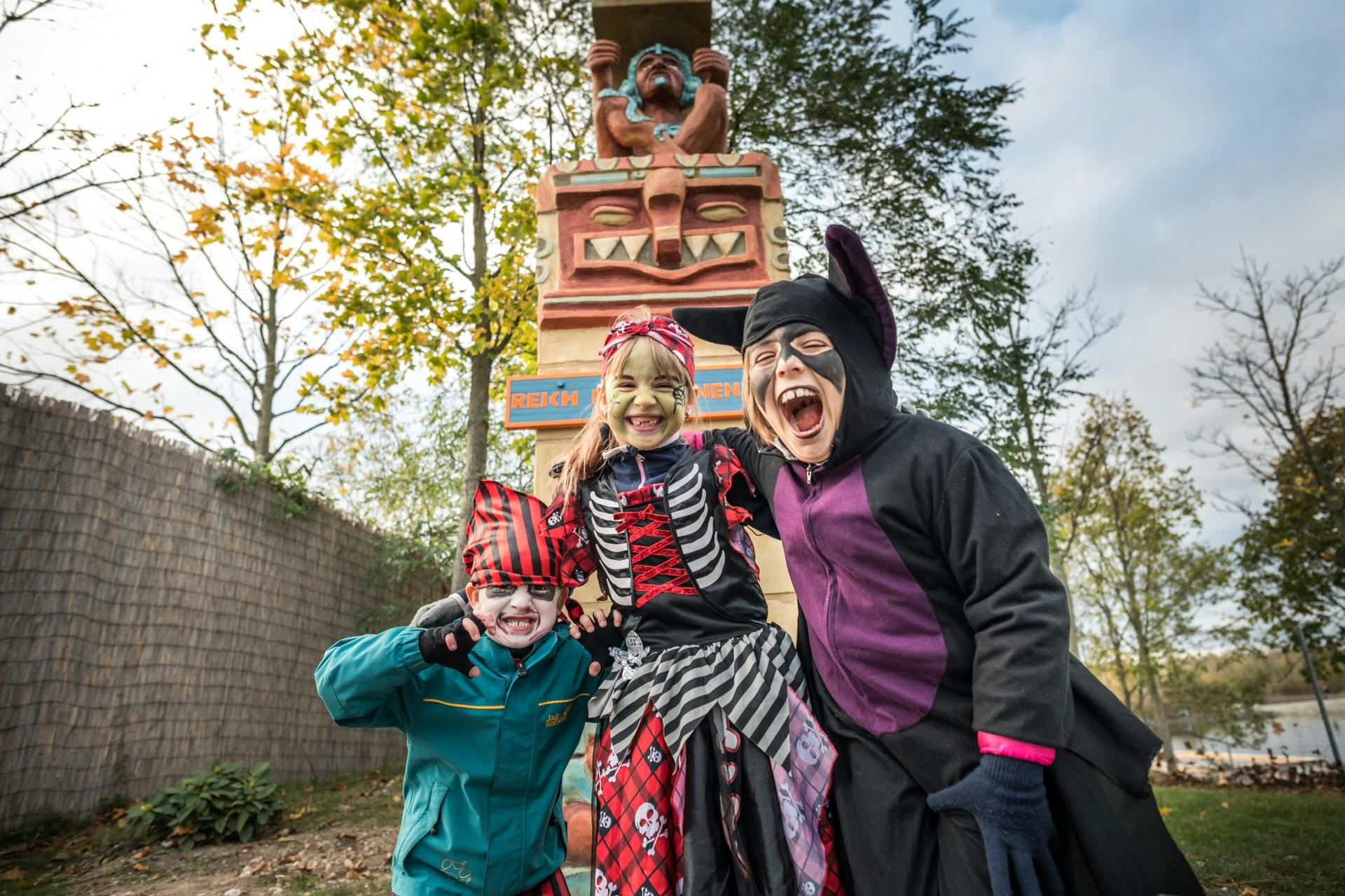 Tre børn i farverige kostumer smiler foran en statue omgivet af træer.