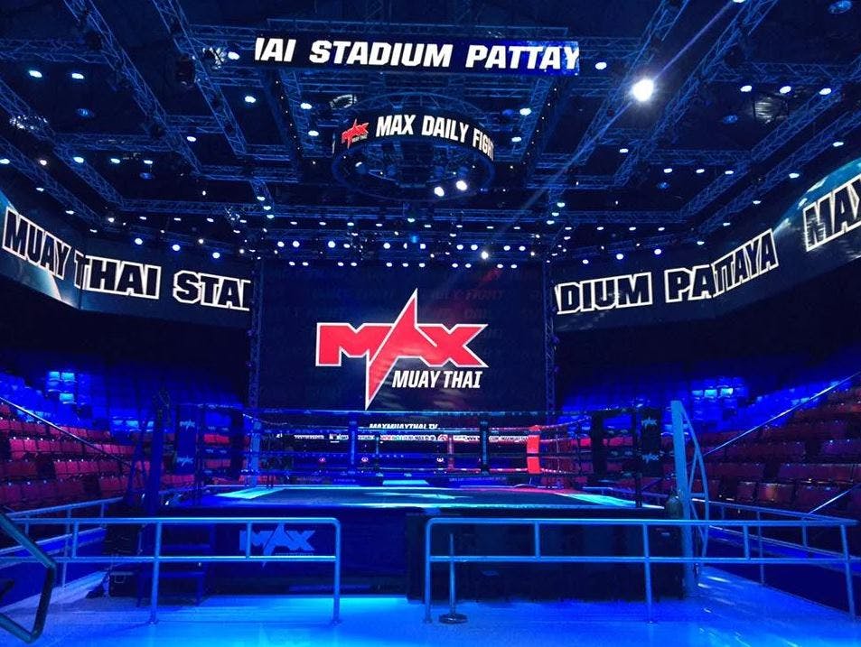 Indendørs Muay Thai-arena med kraftig belysning, store skærme, der viser "MAX Muay Thai", og tomme røde og blå siddepladser.