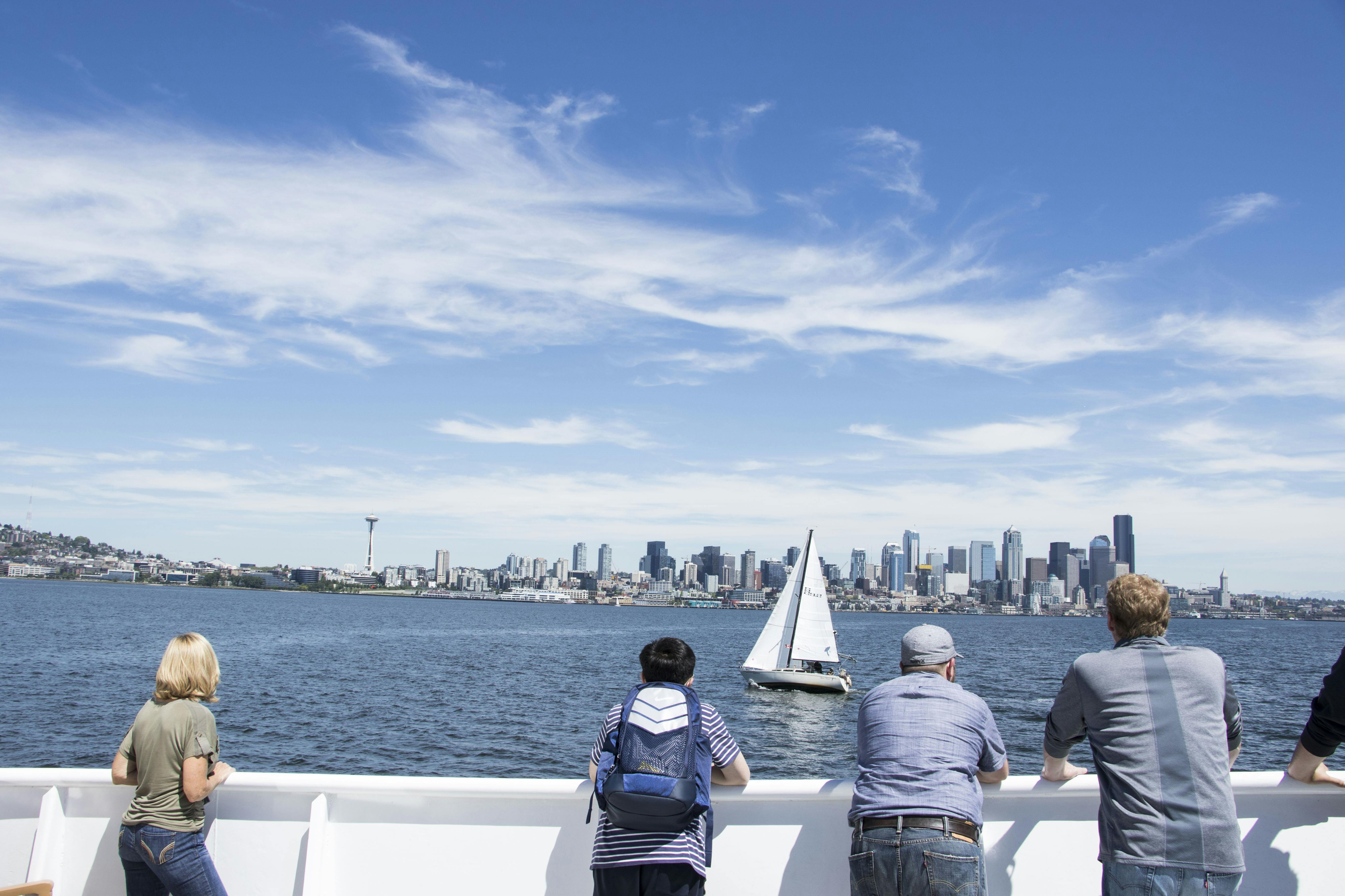 People on a boat look out over water toward a city skyline, with a sailboat in the middle distance under a clear blue sky.