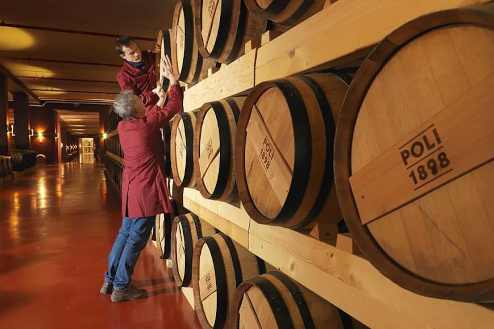 Two people in red coats inspecting large wooden barrels stacked on shelves in a warehouse with a red floor.