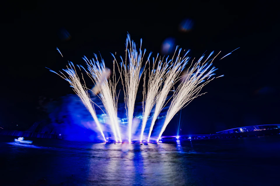 Bright fireworks burst into the night sky above a body of water, illuminated by blue stage lights in the background.