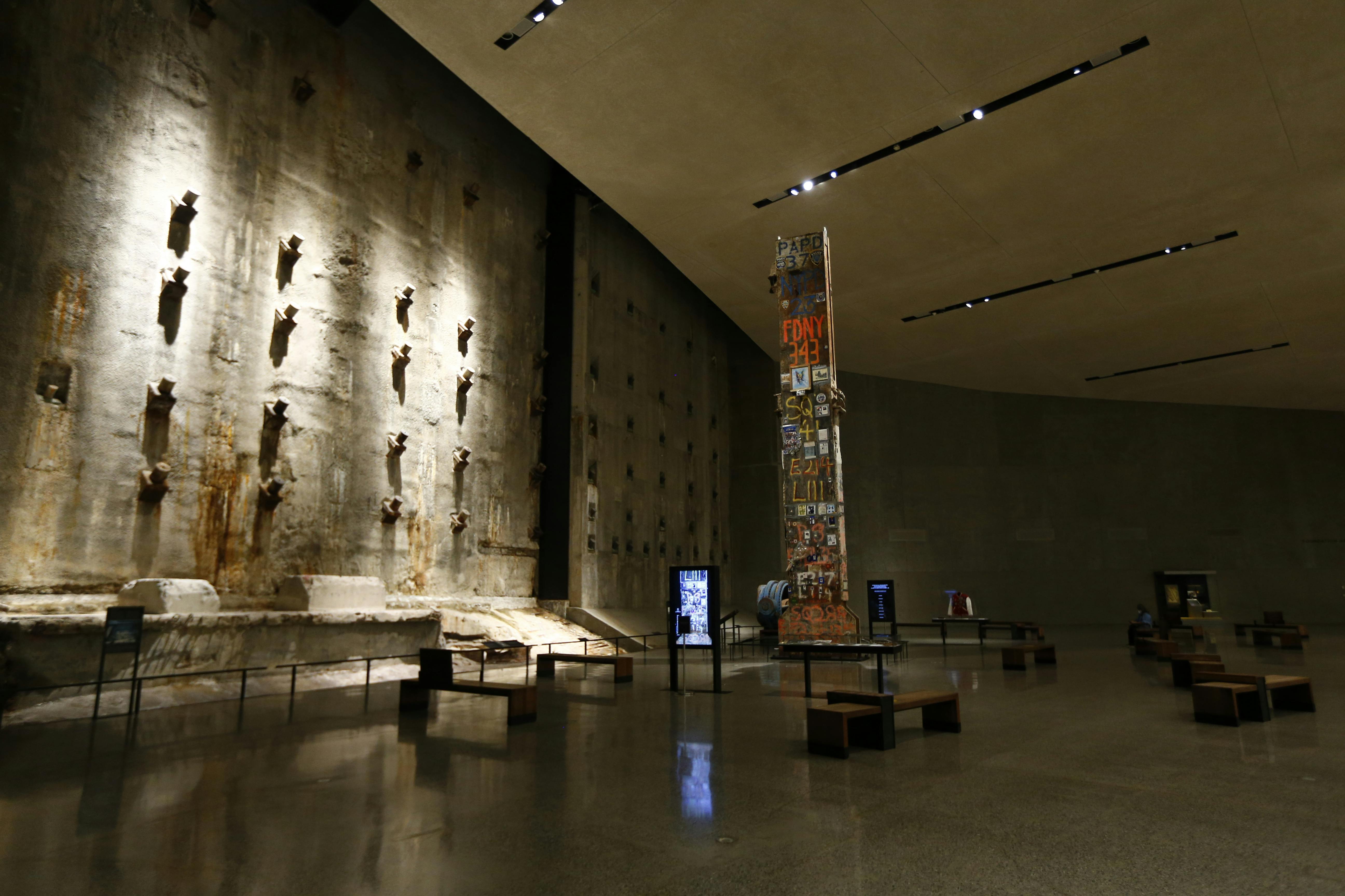 Ευρύχωρη, αμυδρά φωτισμένη αίθουσα μουσείων με μεγάλο τσιμεντένιο τοίχο, στήλη μνήμης καλυμμένη με μηνύματα και αραιά καθίσματα.