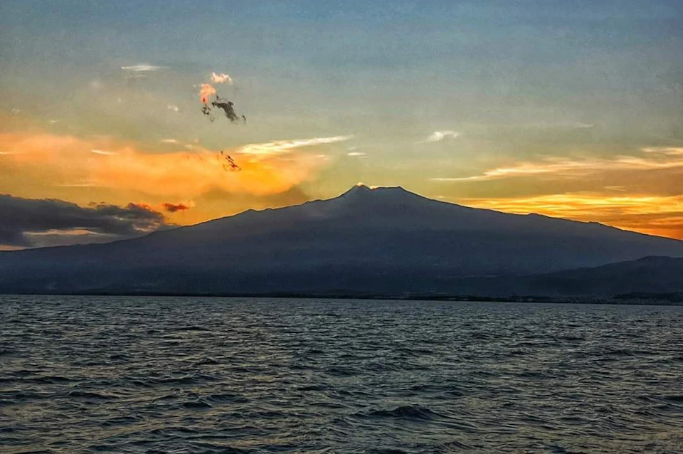A mountain silhouette at sunset with a calm body of water in the foreground and a colorful sky in the background.
