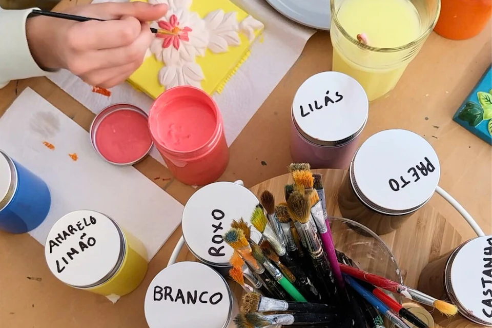 A hand painting a flower on yellow canvas. Paint jars labelled "LILÁS," "ROXO," "BRANCO," and various brushes are visible.