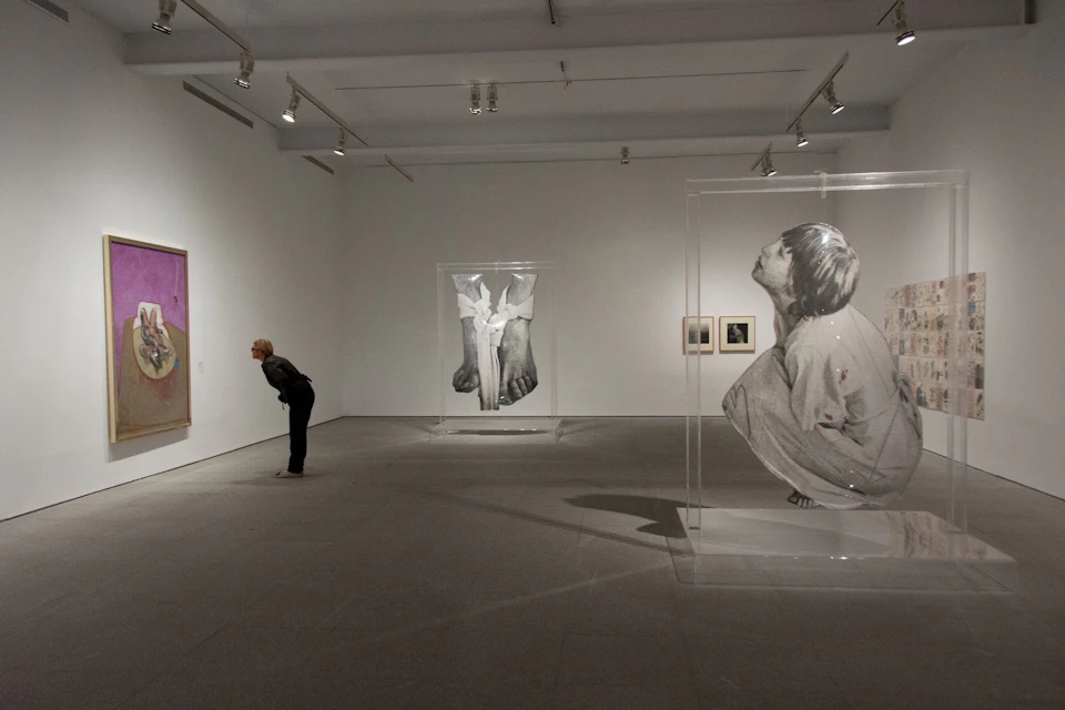 A person leans in to observe artwork in a gallery featuring large-scale grayscale drawings and photographs under bright lighting.