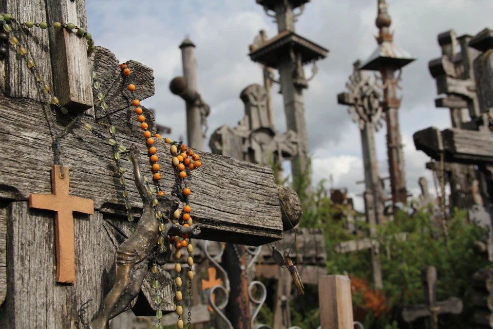 Numerous weathered wooden crosses and rosary beads are seen in an outdoor area under a cloudy sky.