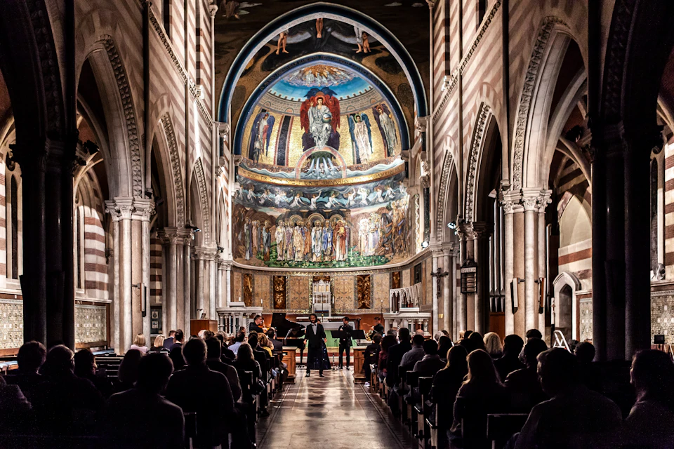 People seated inside a grand cathedral with ornate arches and a detailed fresco above the altar, attending a performance.