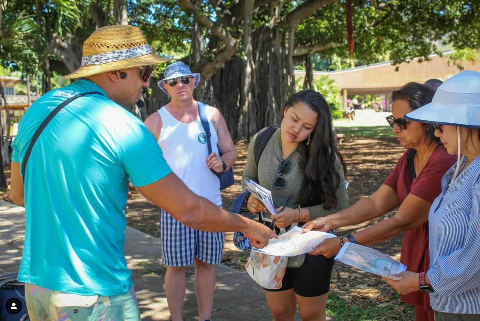 Osoba w słomkowym kapeluszu i niebieskiej koszuli wskazuje na mapę trzymaną przez kobietę. Inna osoba obserwuje w otoczeniu parku.