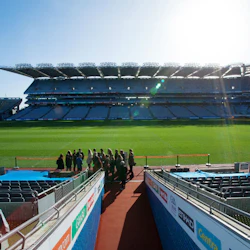 En grupp människor står nära kanten av en stor, tom stadion med ett grönt fält, omgiven av sittplatser.