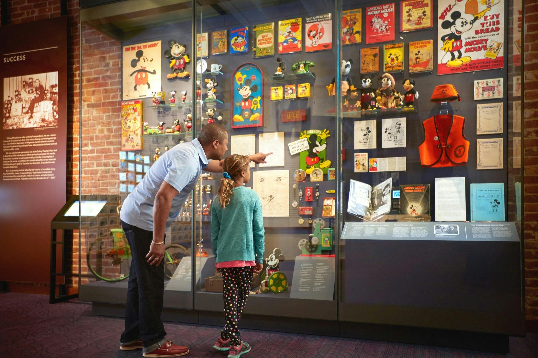 An adult and a child look at a display of vintage Mickey Mouse memorabilia in a glass case at a museum.