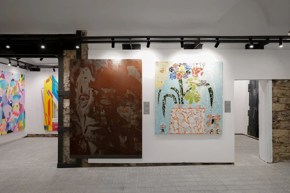 A gallery exhibition featuring two artworks on a wall, one abstract portrait and one floral-themed mosaic.