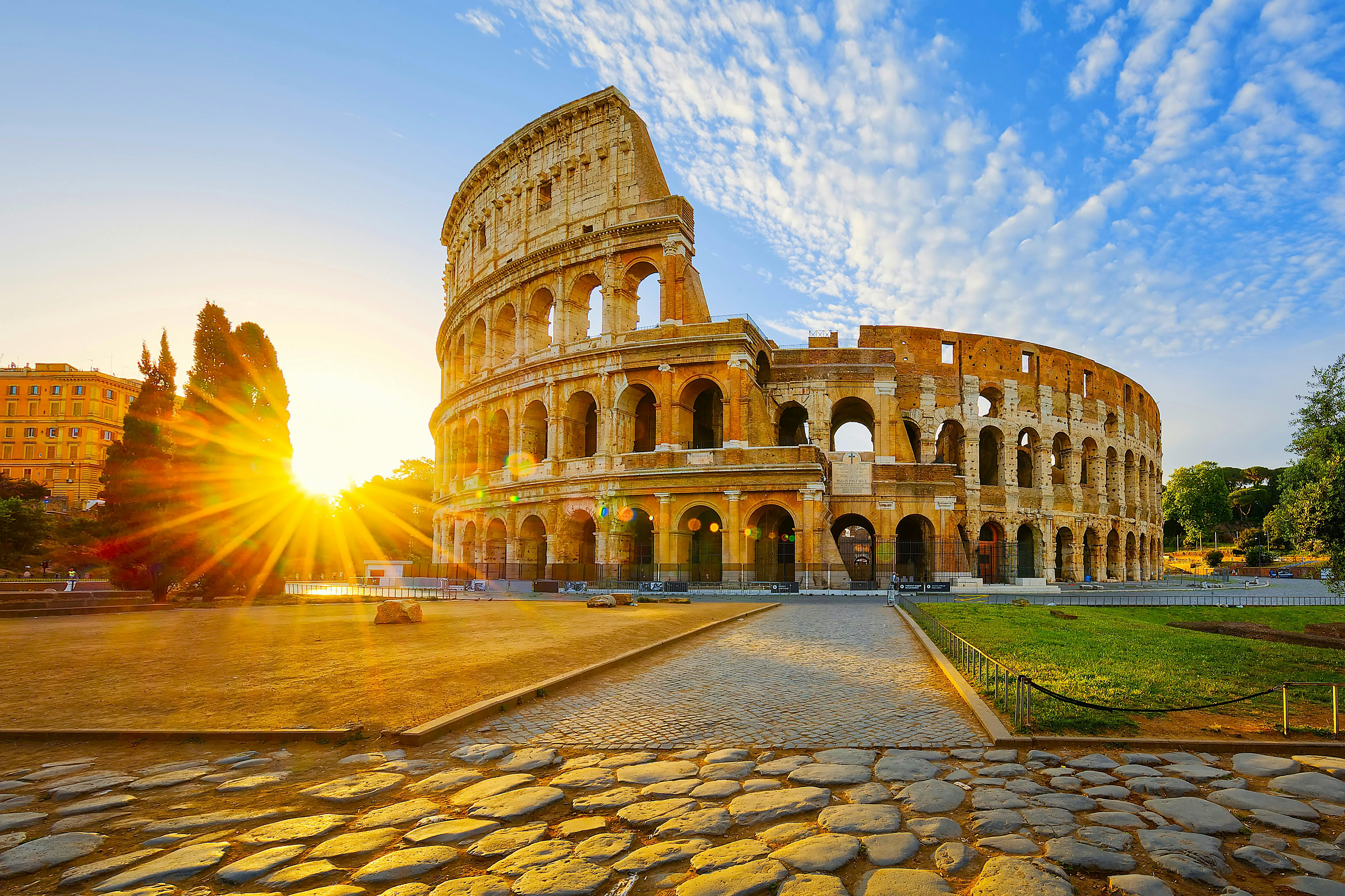 The Colosseum in Rome under a blue sky with scattered clouds, sunlight shining from the left, and a cobblestone path in front.