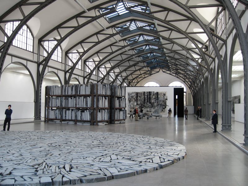 Interiér v industriálním stylu s klenutými ocelovými nosníky, kruhovou kamennou instalací, vysokou regálovou konstrukcí a návštěvníky v pozadí.