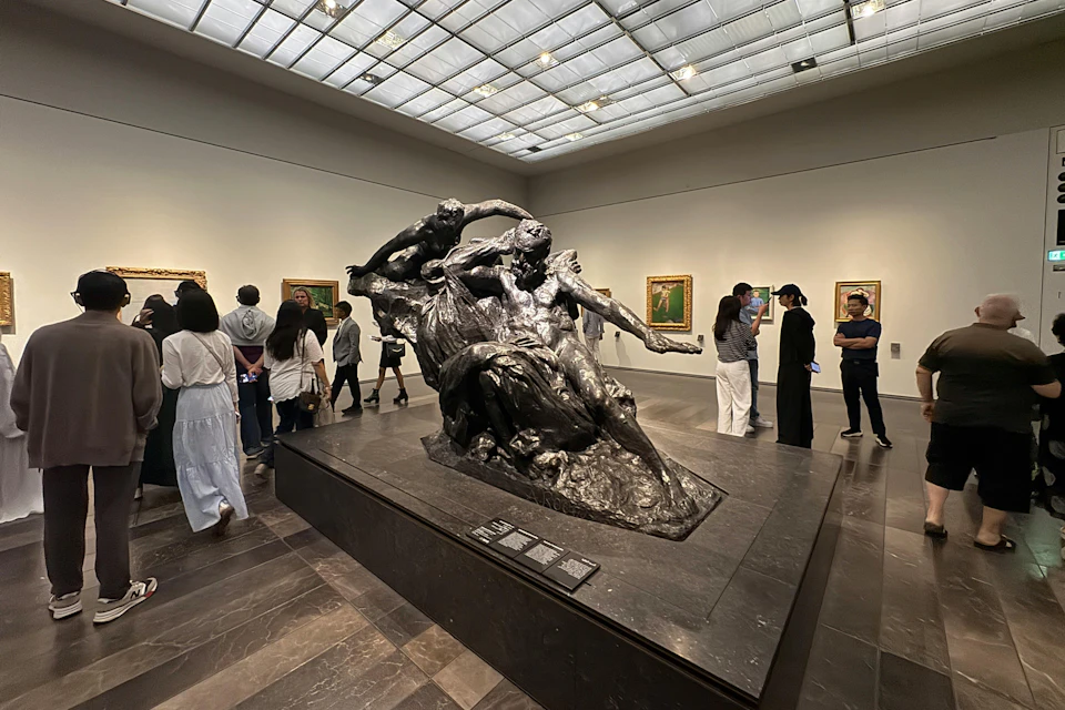 Museum gallery with a bronze sculpture as the centerpiece, surrounded by paintings on the walls and visitors observing the art.