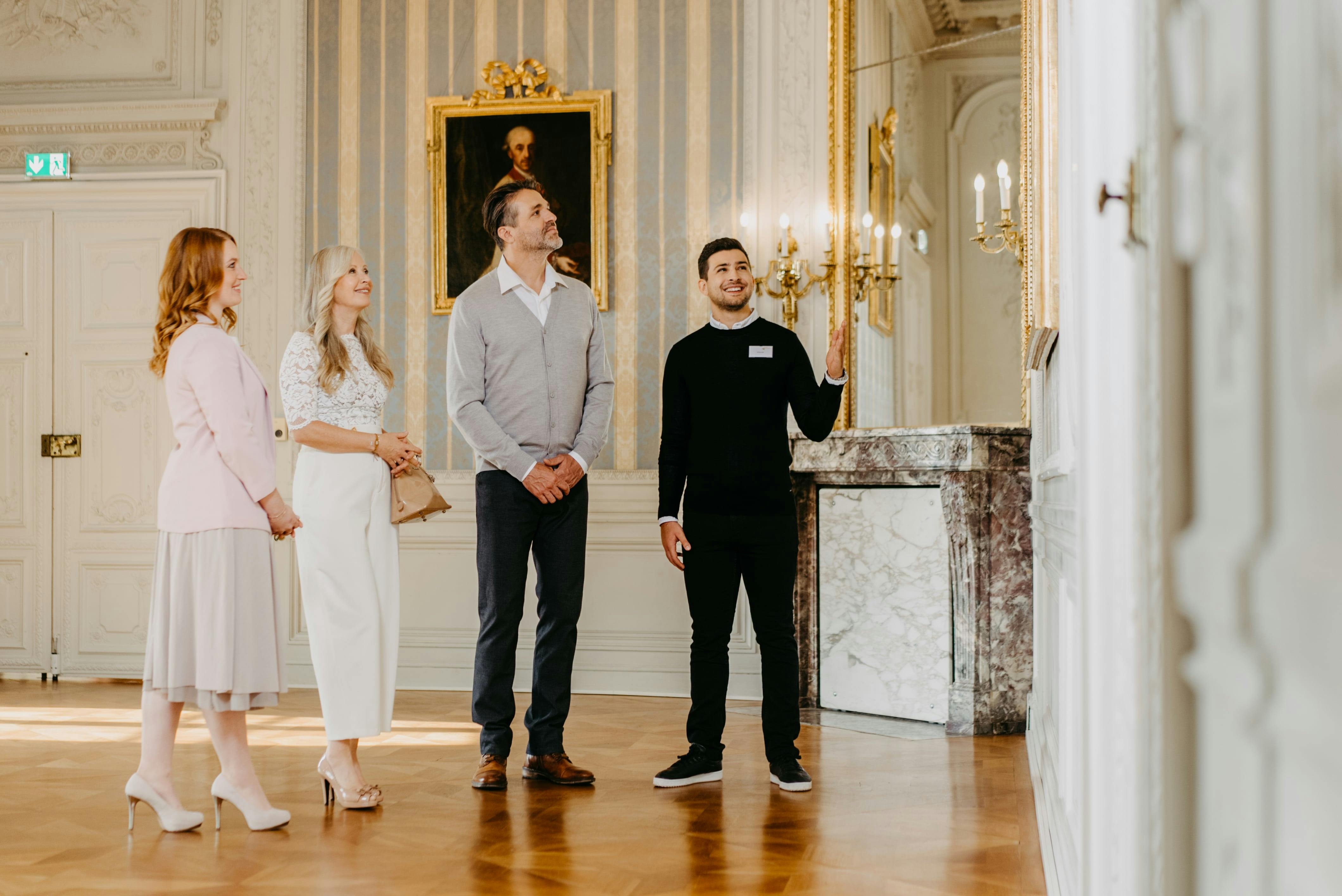 Ένας ξεναγός κάνει χειρονομίες ενώ μιλάει σε τρεις ενήλικες σε ένα κομψό δωμάτιο με πορτρέτο και πολυέλαιο.