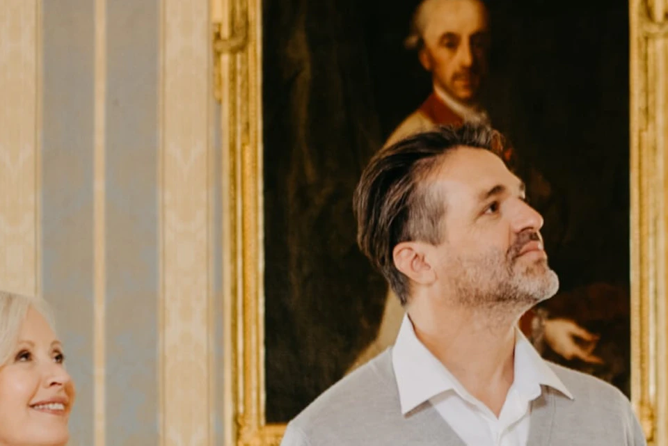 A tour guide gestures while speaking to three adults in an elegant room with a portrait and chandelier.