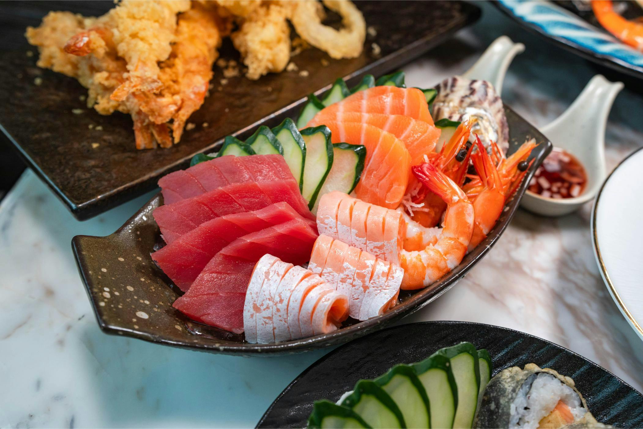 A plate of assorted sushi and sashimi, featuring tuna, salmon, shrimp, cucumber slices, and tempura in the background.