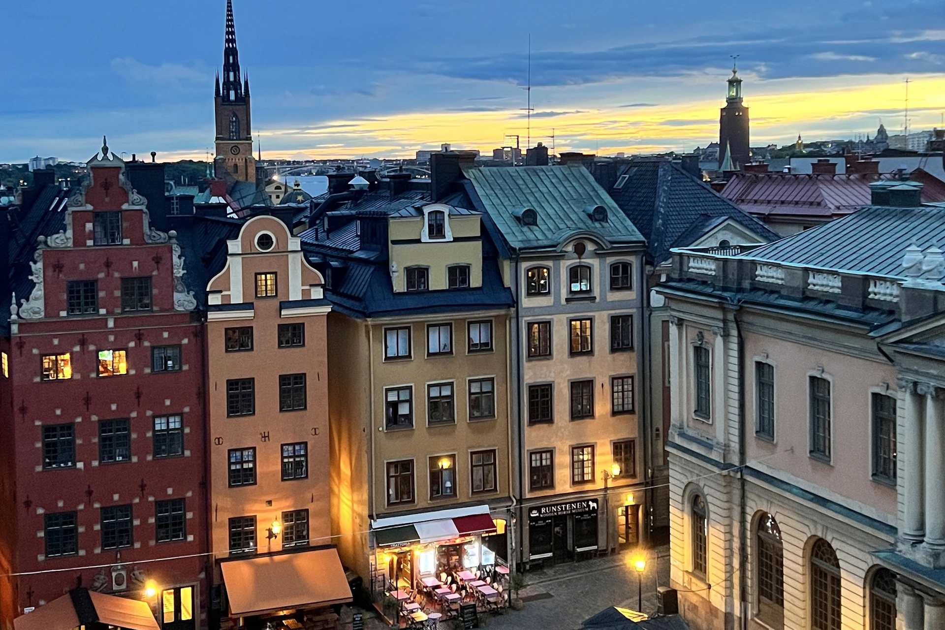 Evening view of a charming European street with colorful buildings, a small outdoor café, and distant church steeples under a glowing sky.