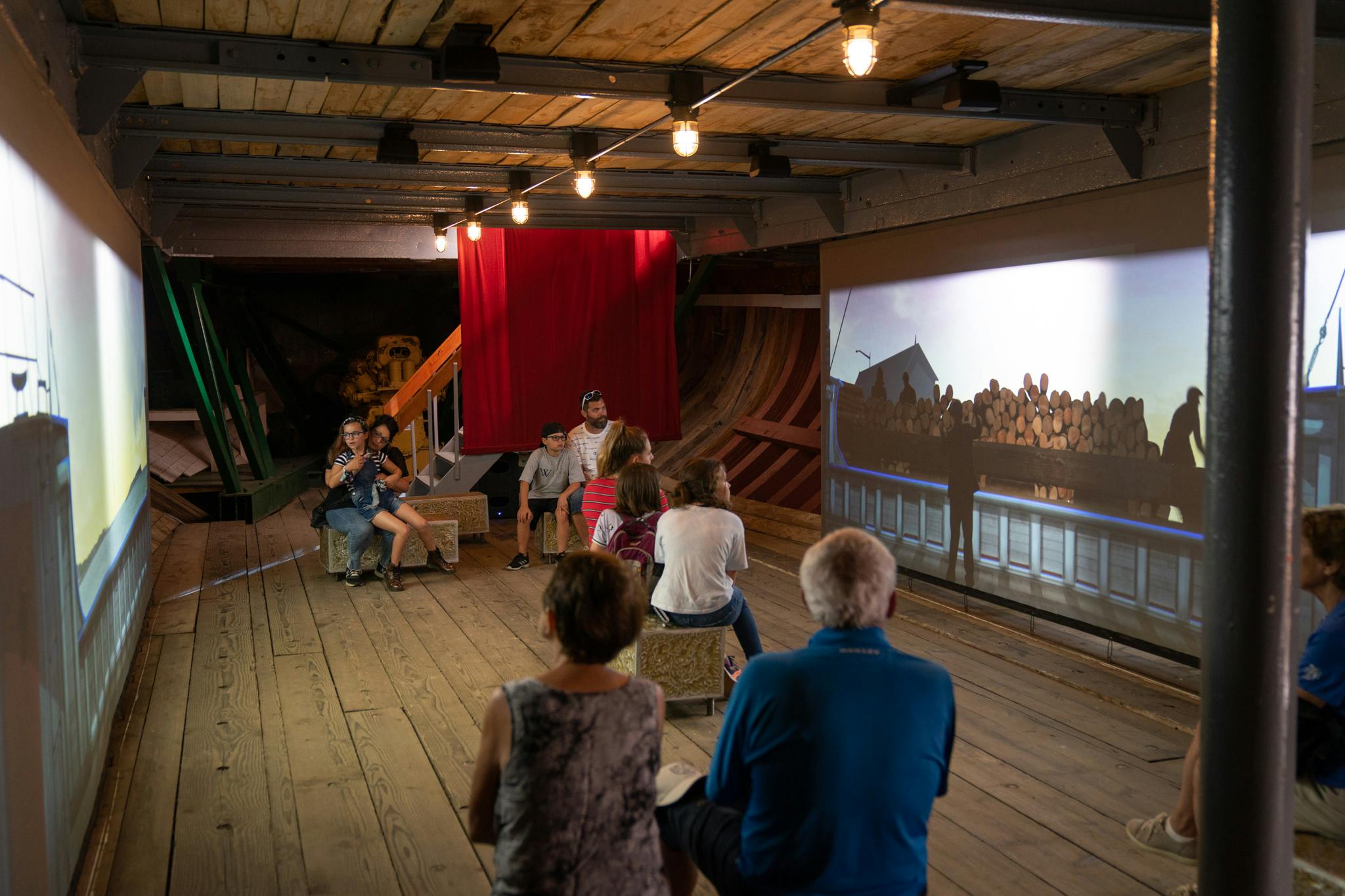 Folk, der sidder på taburetter i en træmuseumsudstilling, ser en skærm, der viser en person, der står ved stablede træstammer.