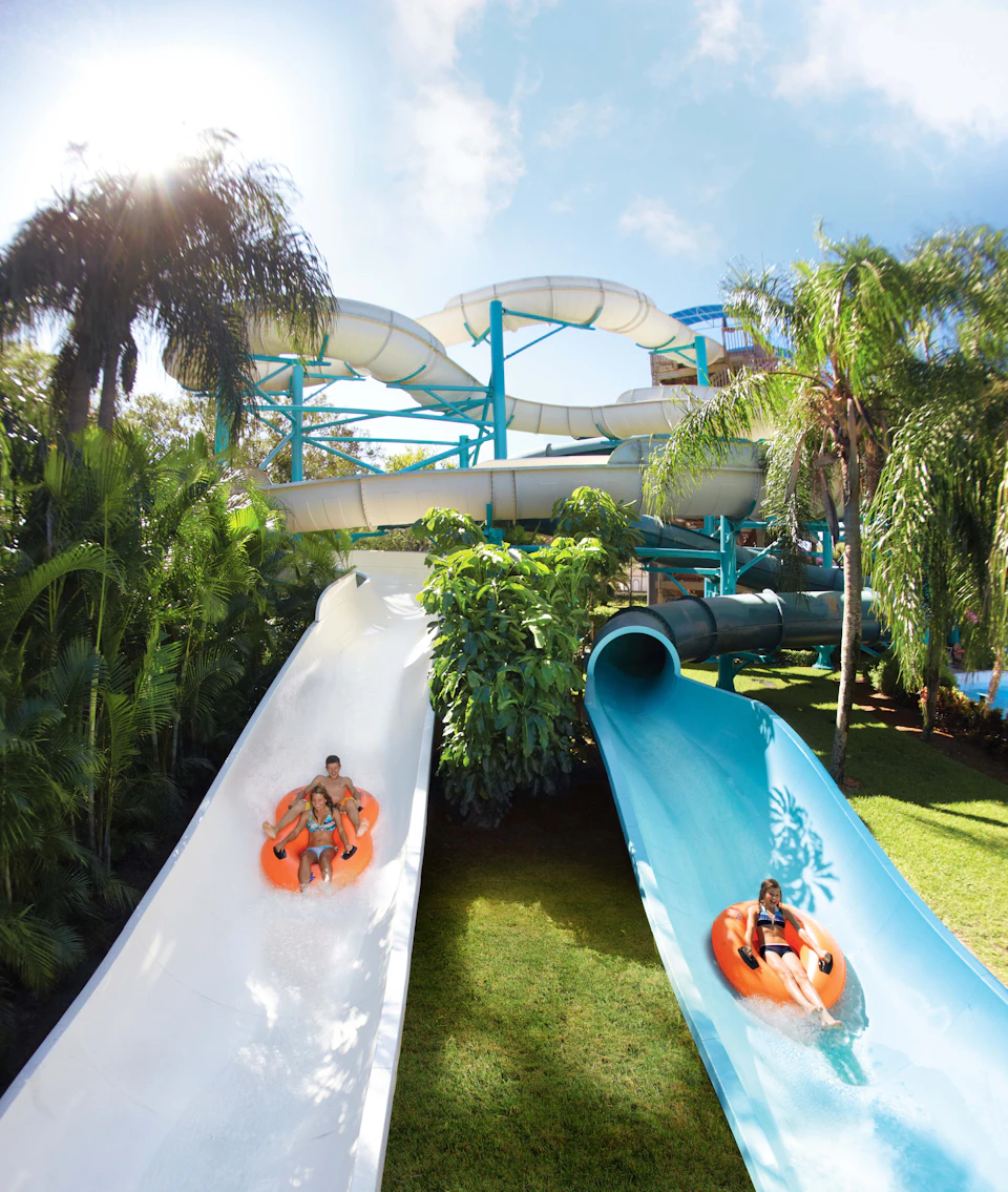 Two people going down a white waterslide on an orange raft beside another person on a blue waterslide, surrounded by lush greenery.