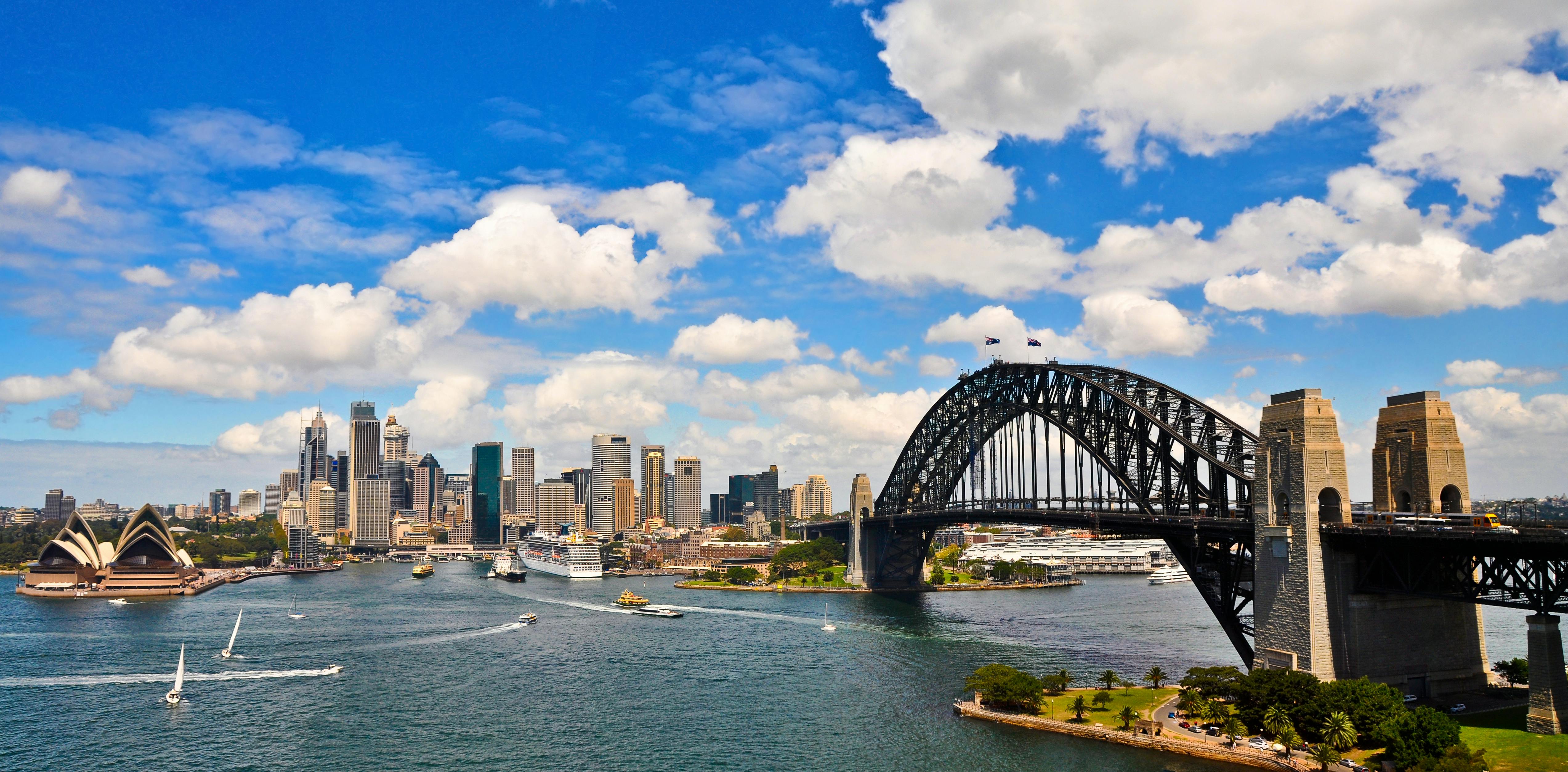 The image shows a city skyline with a prominent bridge, opera house, and various boats in the water under a partly cloudy sky.