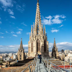 Människor står på en metallgång framför en katedralspiral i gotisk stil mot en ljusblå himmel med spridda moln.