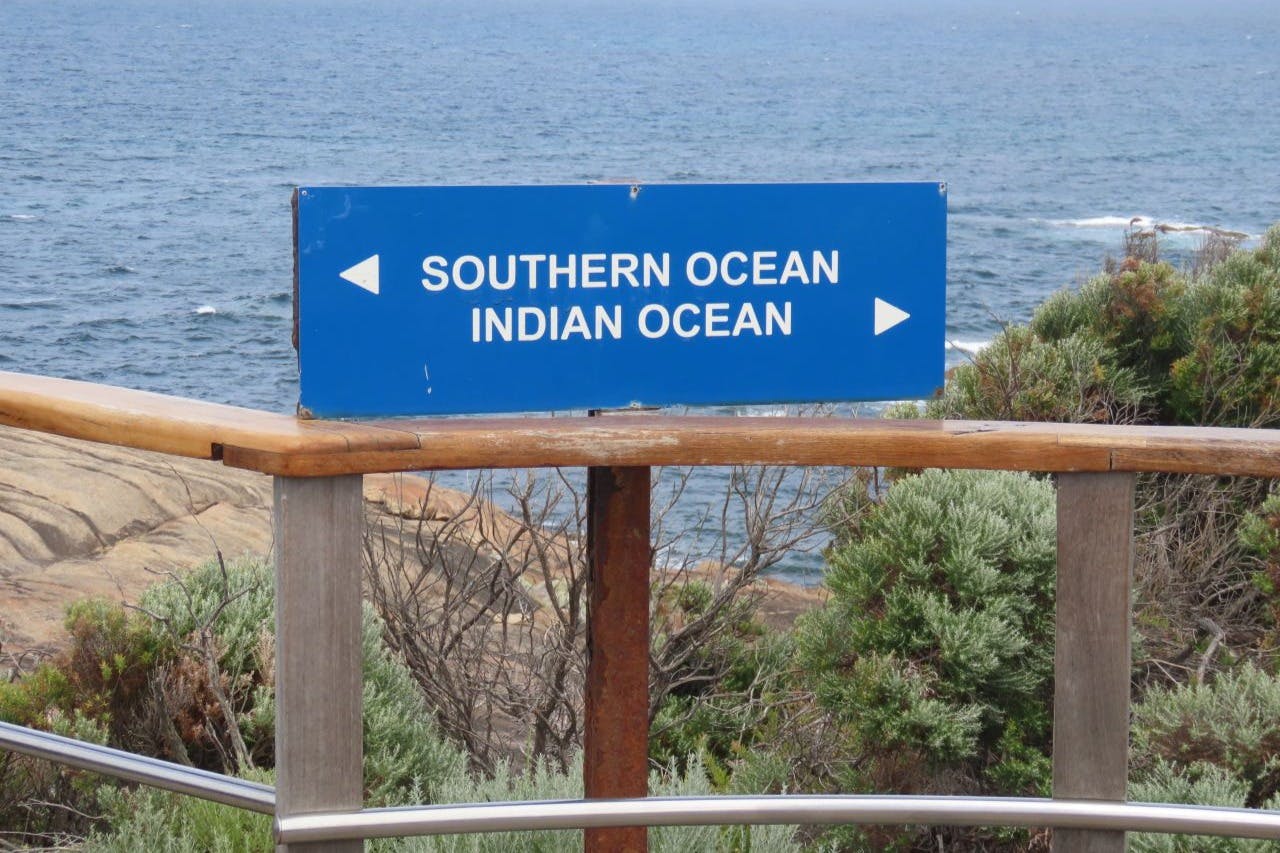 Niebieski znak kierunkowy z napisem „Ocean Południowy” i „Ocean Indyjski” ze strzałkami, umieszczony w pobliżu obszaru przybrzeżnego z roślinnością.