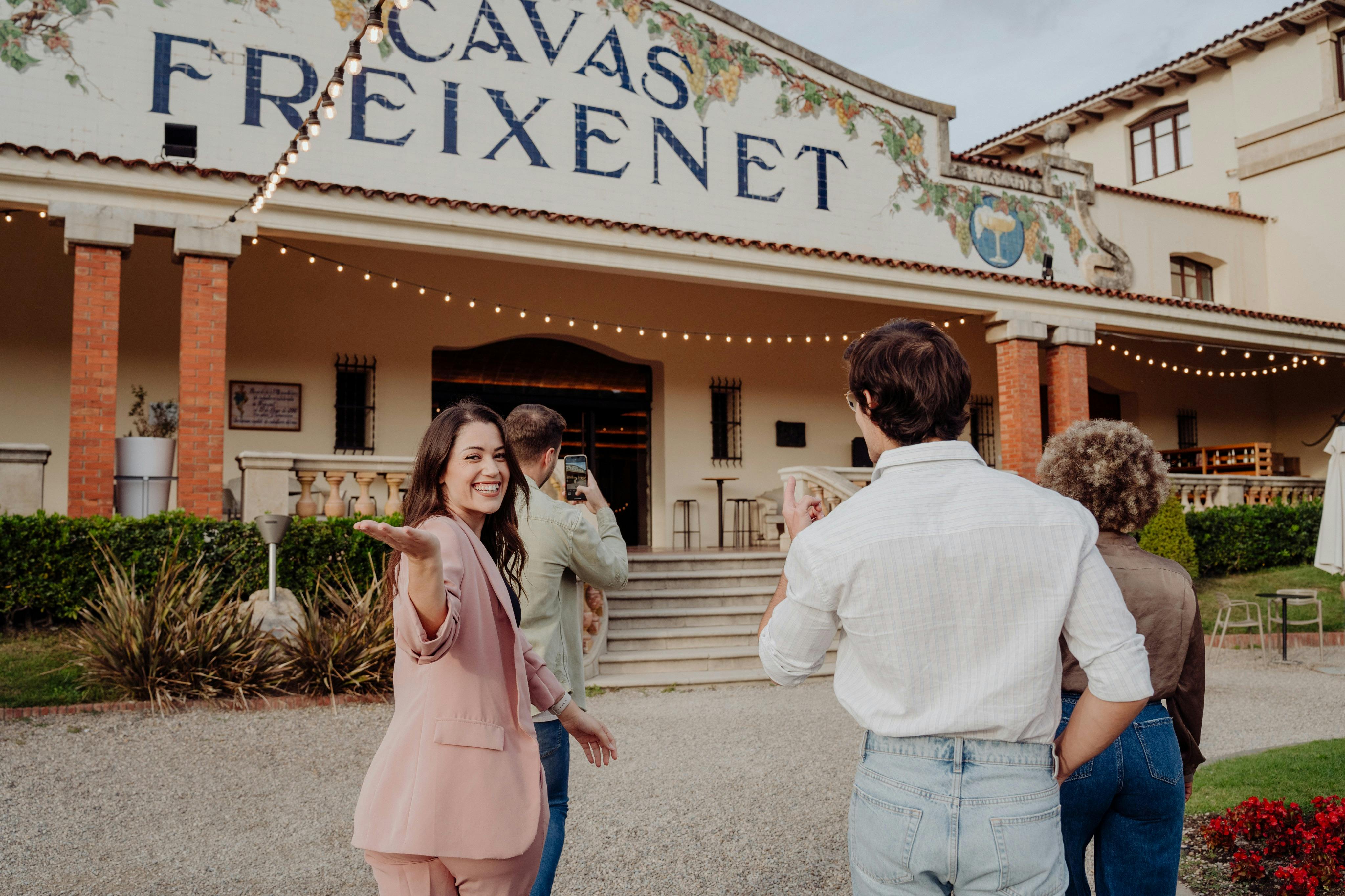 A woman in a pink suit smiles and gestures towards a building labeled "Cavas Freixenet," with two other people nearby.