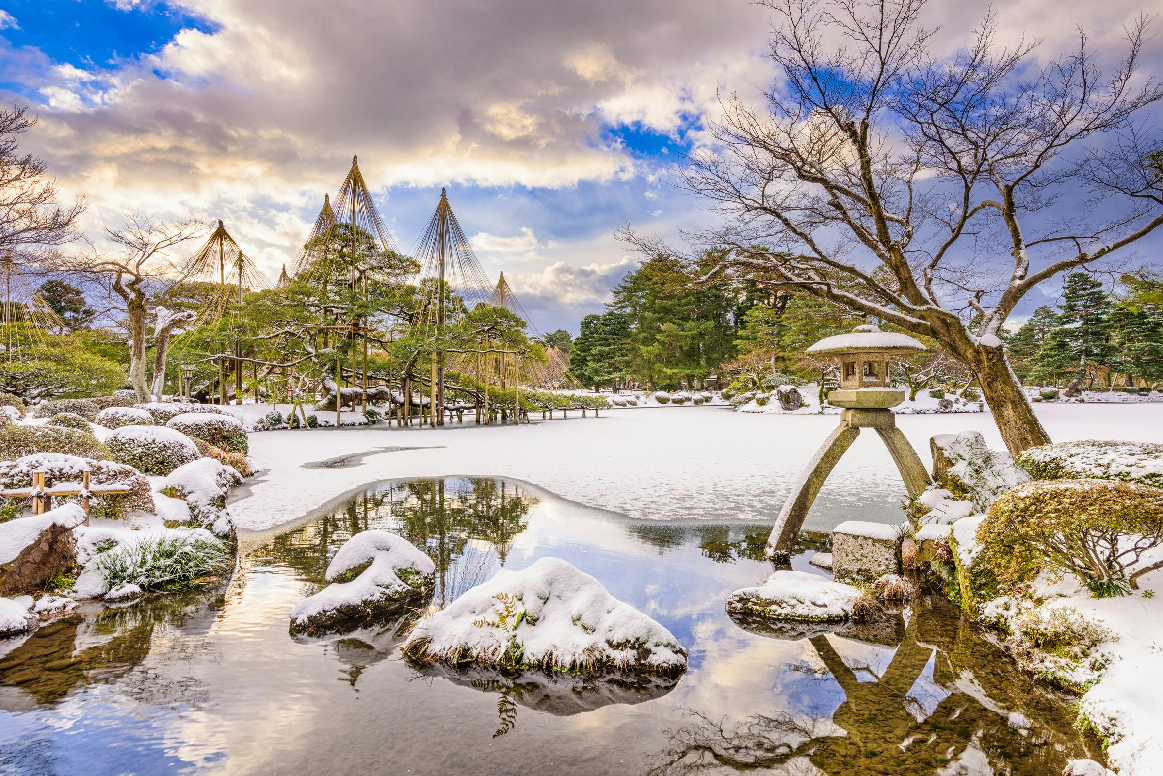 A serene Japanese garden in winter with snow-covered trees, a pond with rocks, and a stone lantern under a cloudy sky.