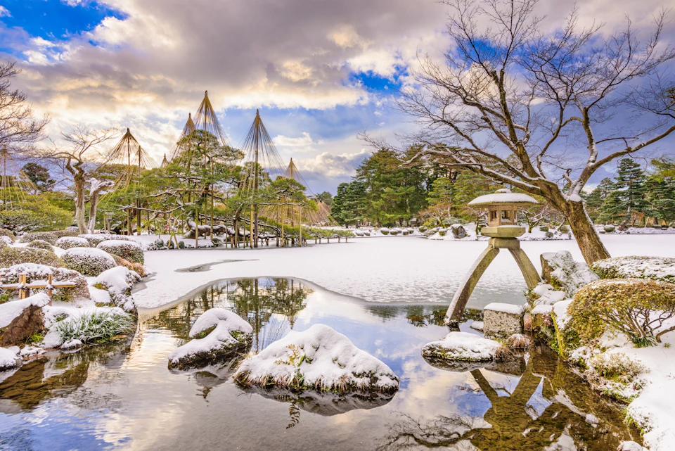 A serene Japanese garden in winter with snow-covered trees, a pond with rocks, and a stone lantern under a cloudy sky.