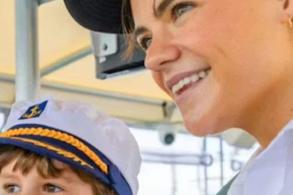 A woman and a child wearing sailor hats are at the helm of a boat, smiling, with water and a cityscape in the background.