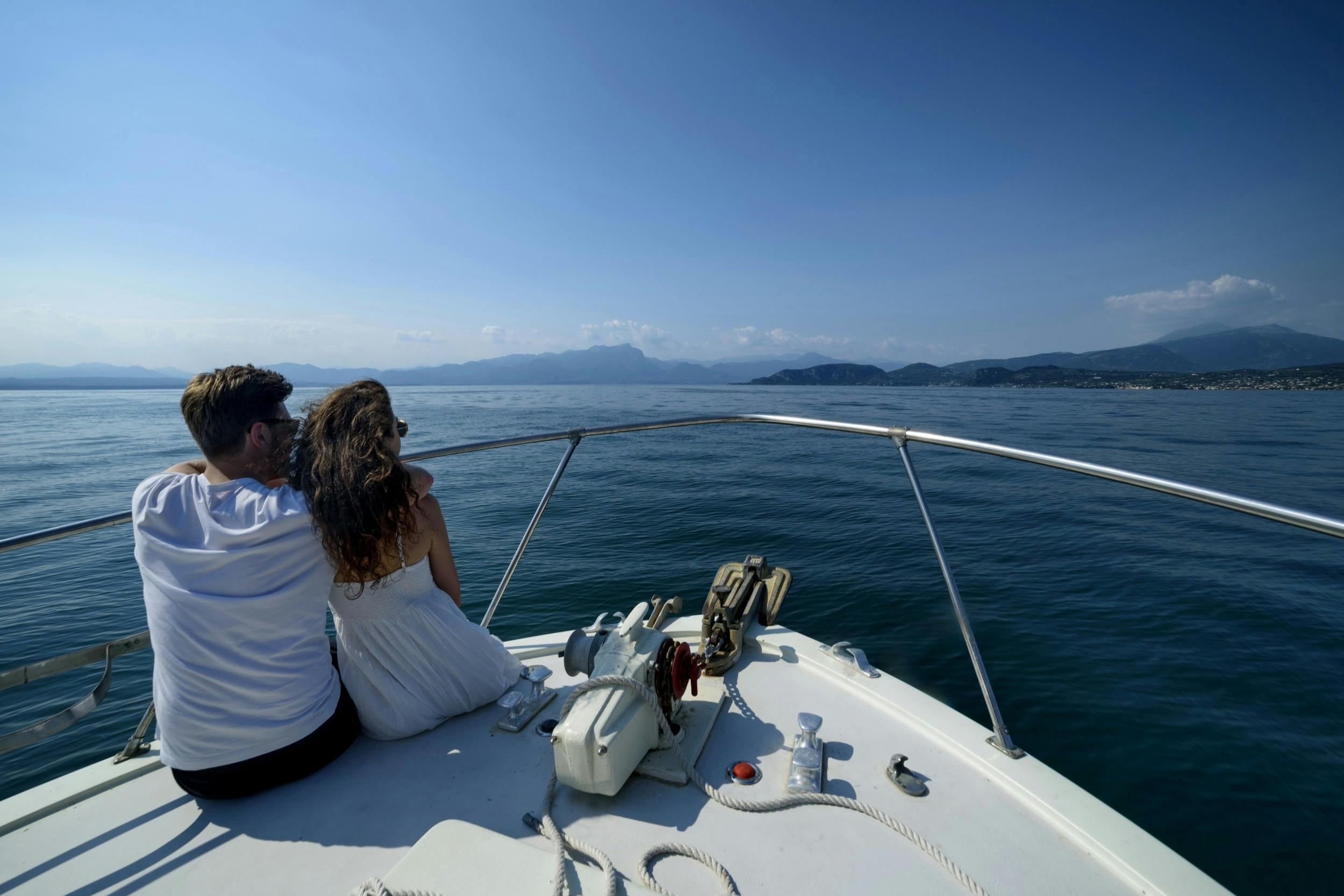 A couple sits at the bow of a boat on a calm sea, looking towards distant mountains under a clear blue sky.