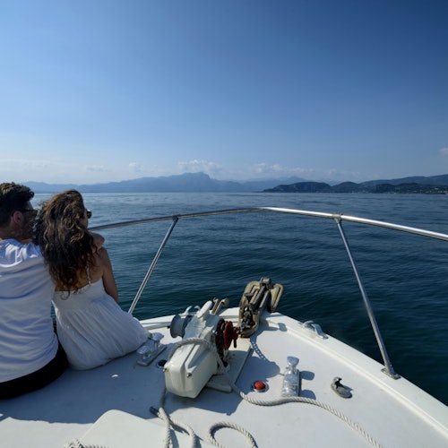 A couple sits at the bow of a boat on a calm sea, looking towards distant mountains under a clear blue sky.