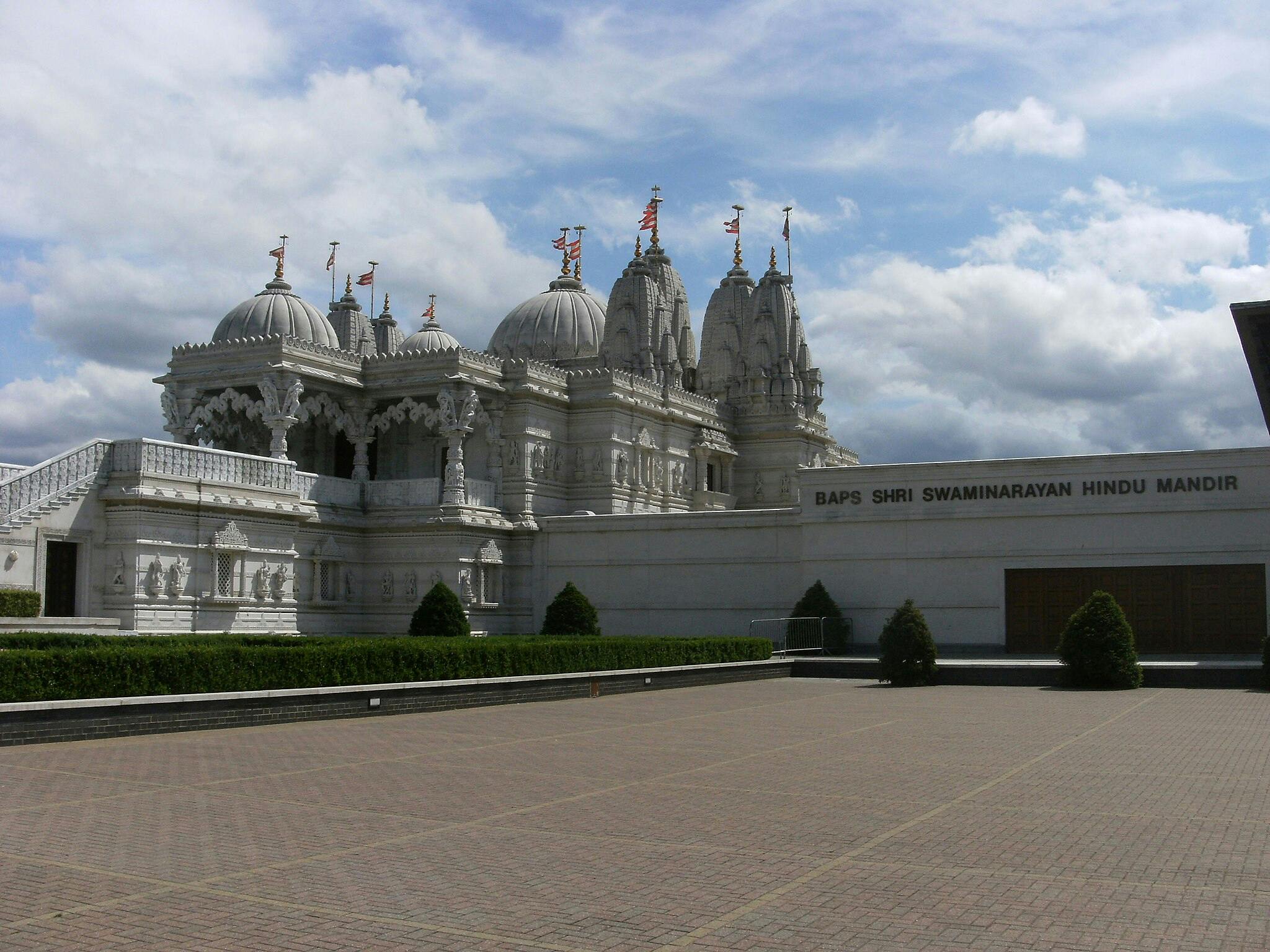 Temple hindou blanc sculpté avec des dômes et des flèches, panneau indiquant "BAPS Shri Swaminarayan Hindu Mandir", sous un ciel nuageux.