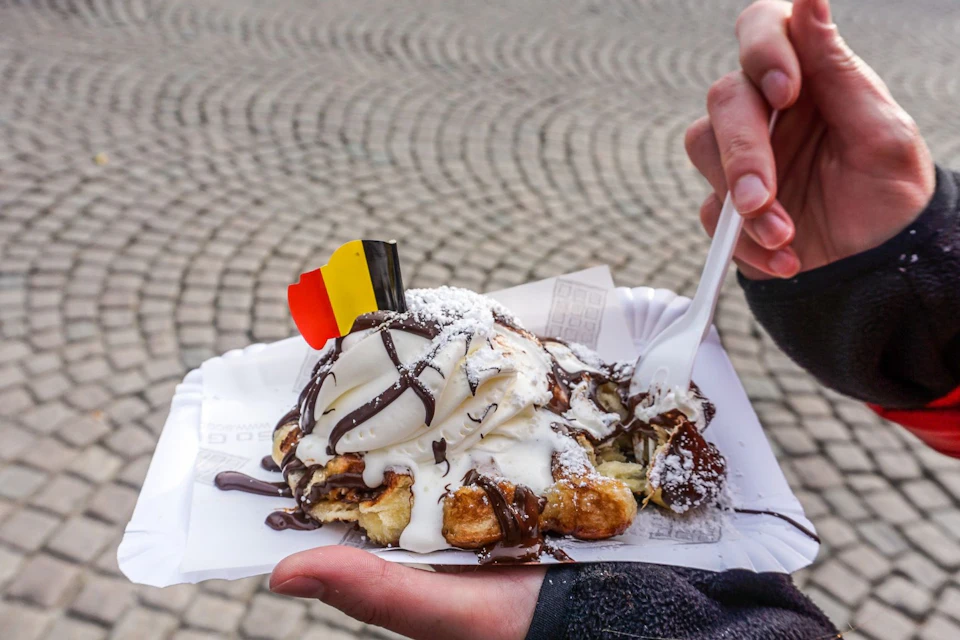 Hand holding a paper plate with a waffle topped with ice cream, chocolate sauce, powdered sugar, and a small Belgian flag.