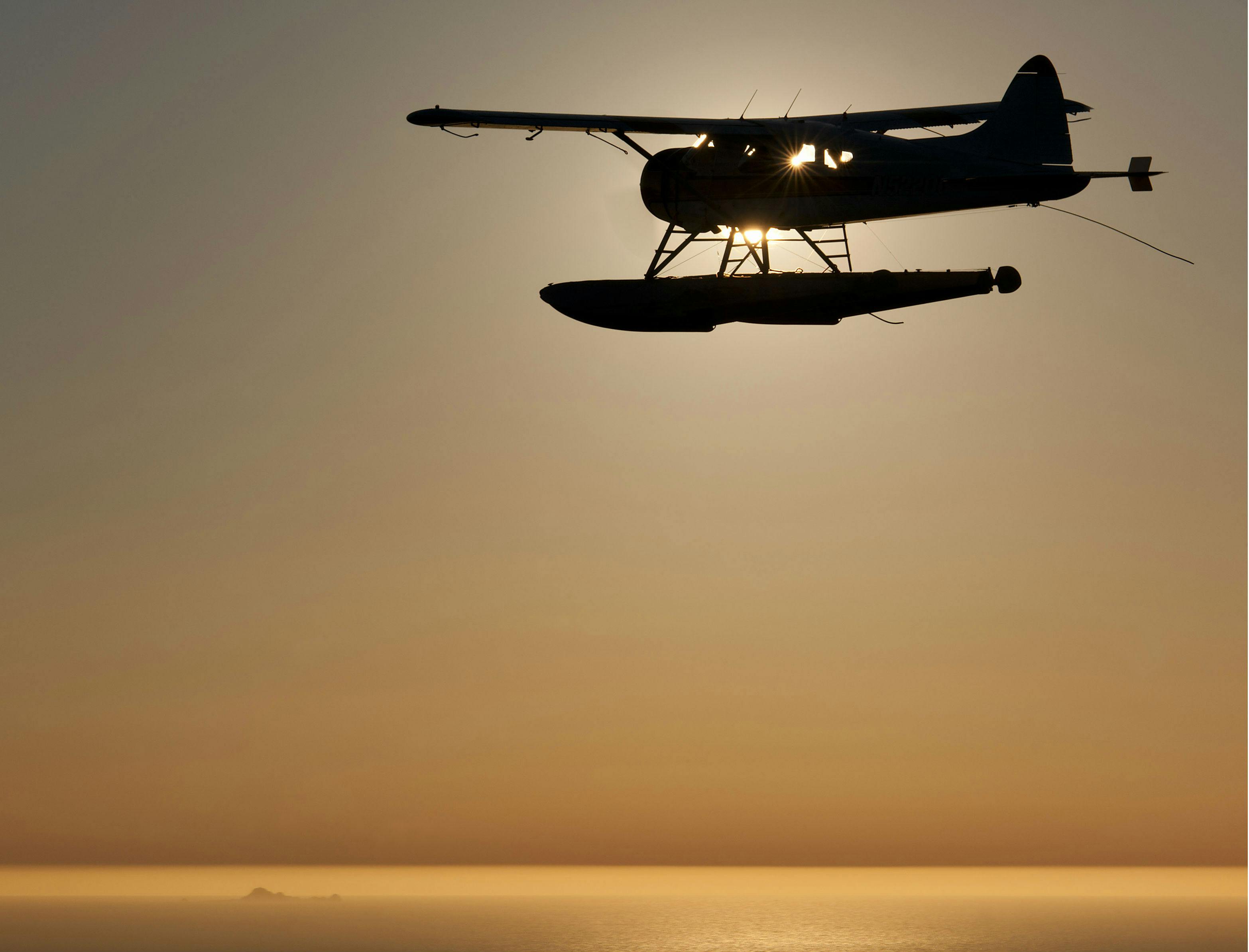 A silhouetted seaplane flying over calm water at sunset with the sun shining through its windows.