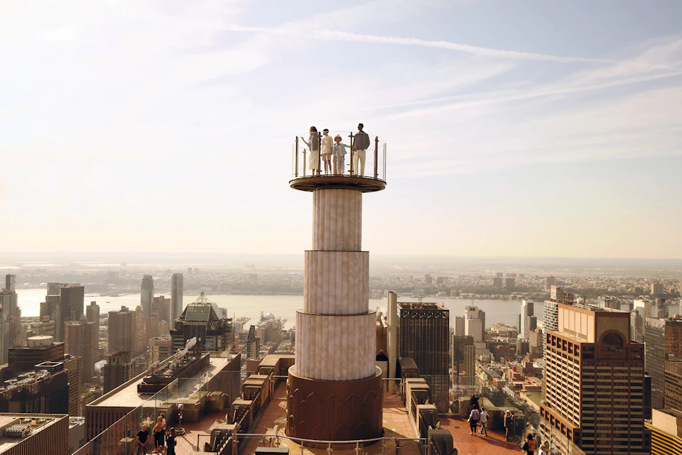 Several people stand on a high tower observation deck overlooking a city skyline with tall buildings and a river.