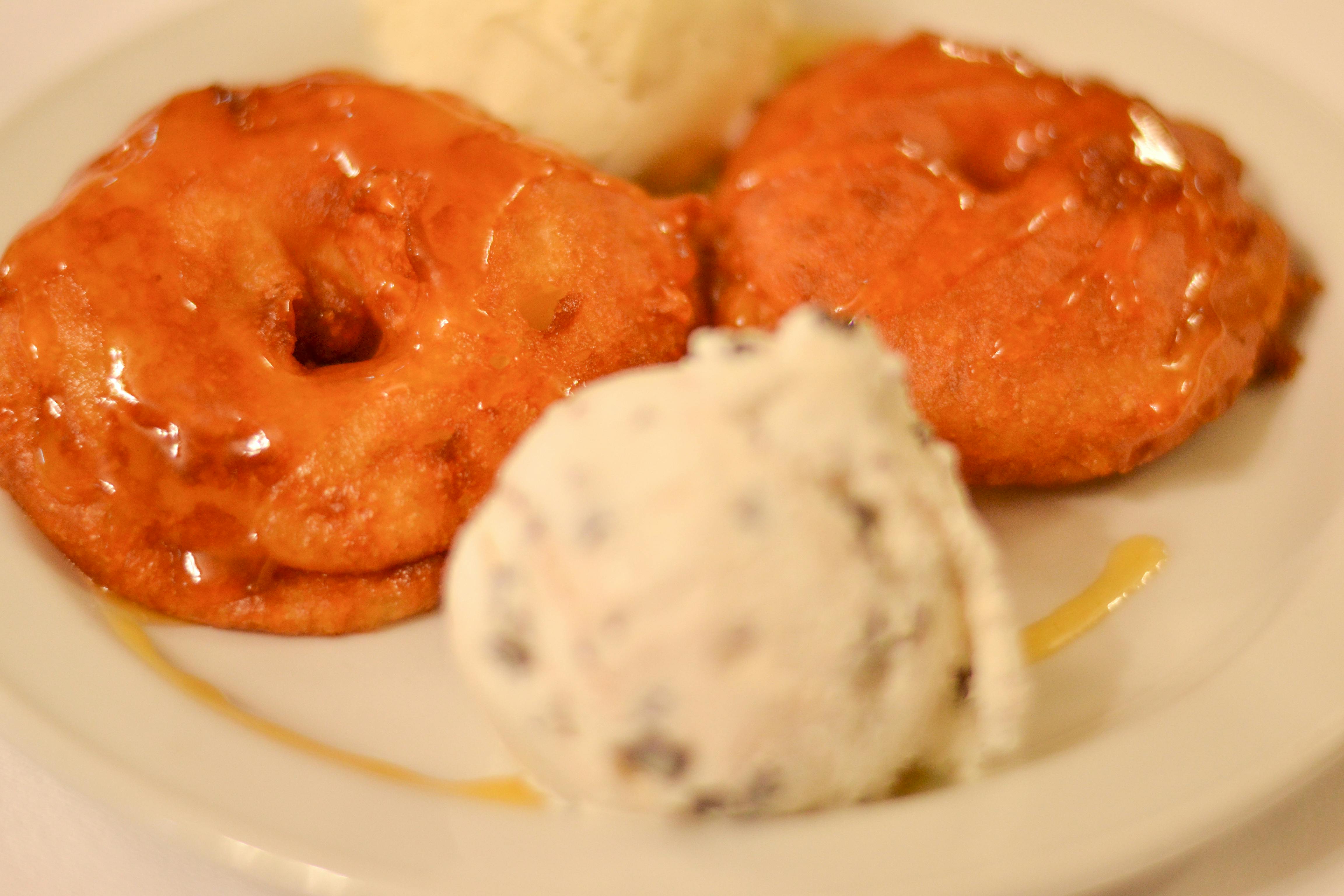 Zwei goldbraune Krapfen mit Sirup beträufelt, begleitet von Kugeln Vanille- und Schokoladeneis auf einem weißen Teller.