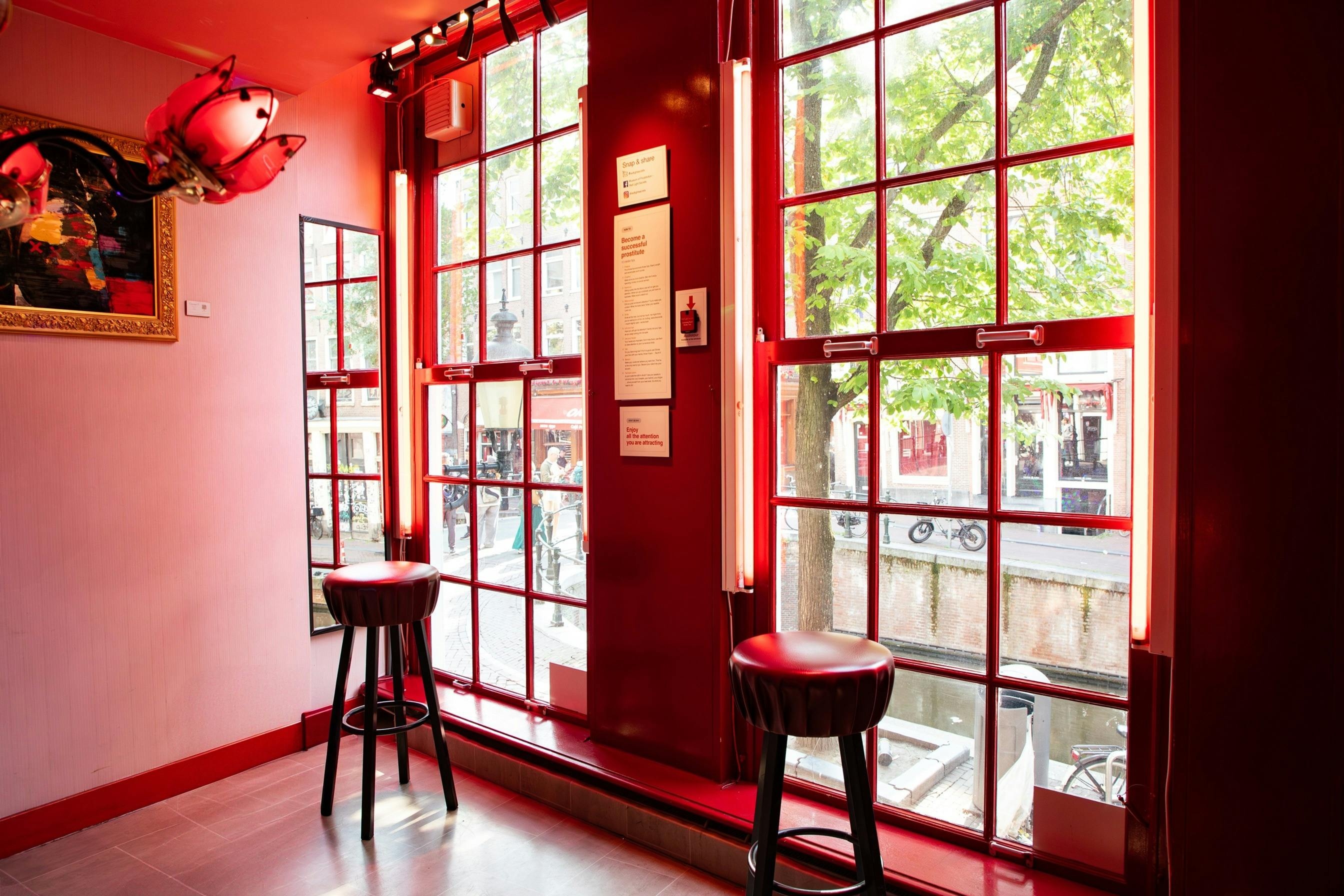 Due sgabelli si trovano accanto a grandi finestre con cornici rosse che si affacciano su un canale e sugli alberi, con specchi e opere d'arte a parete in un interno molto illuminato.