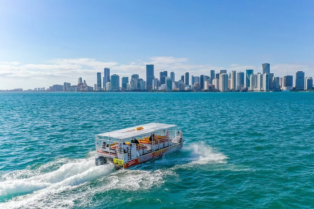 A boat cruising in blue water with a city skyline of tall buildings under a clear blue sky in the background.