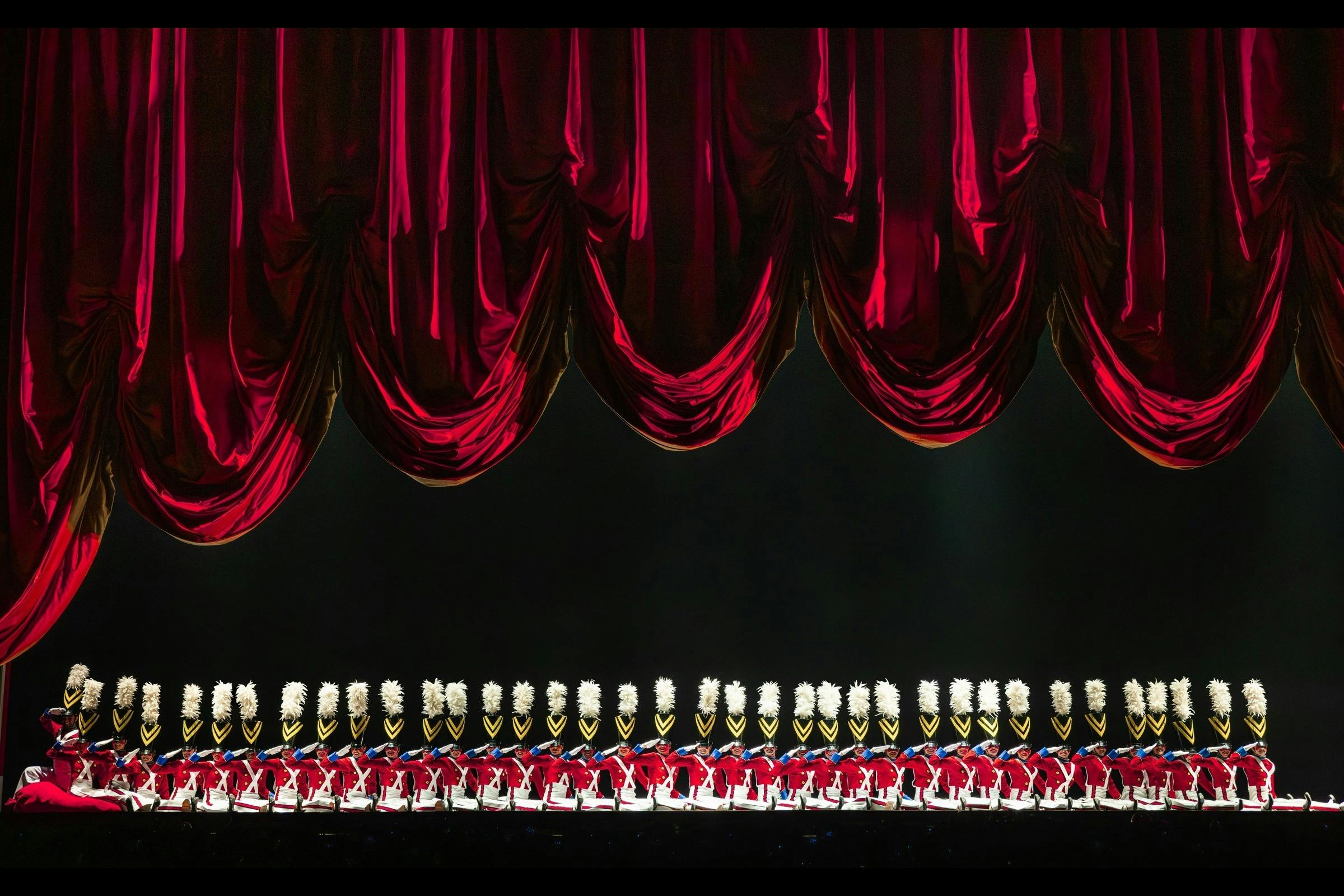 A line of performers dressed as toy soldiers in red uniforms and white hats stands under red stage curtains.