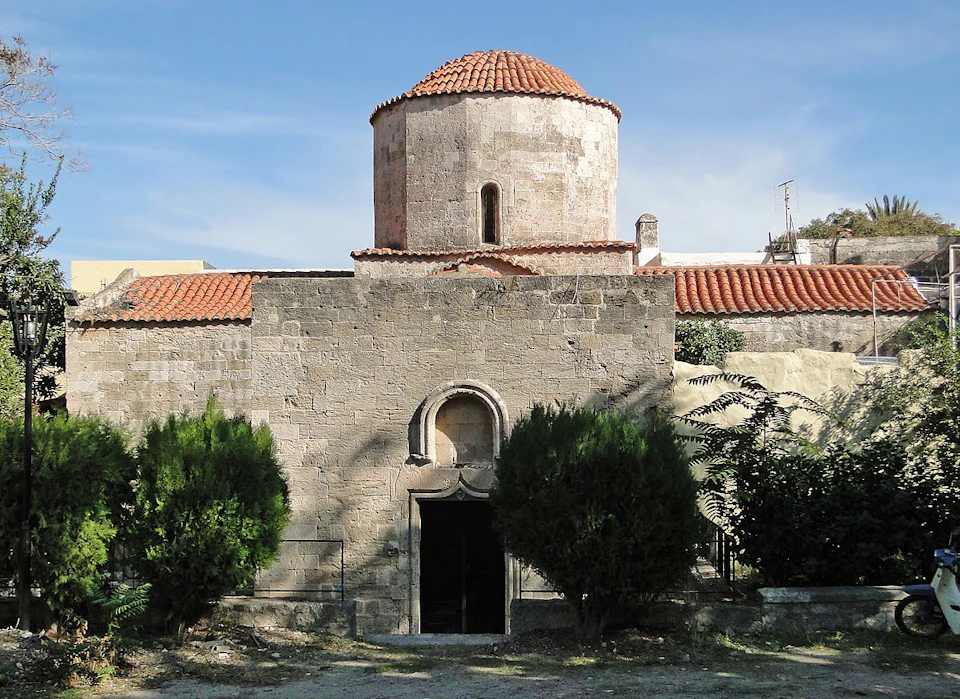 Stone building with a red dome roof, arched entrance, and flanked by two green shrubs, under a clear blue sky.