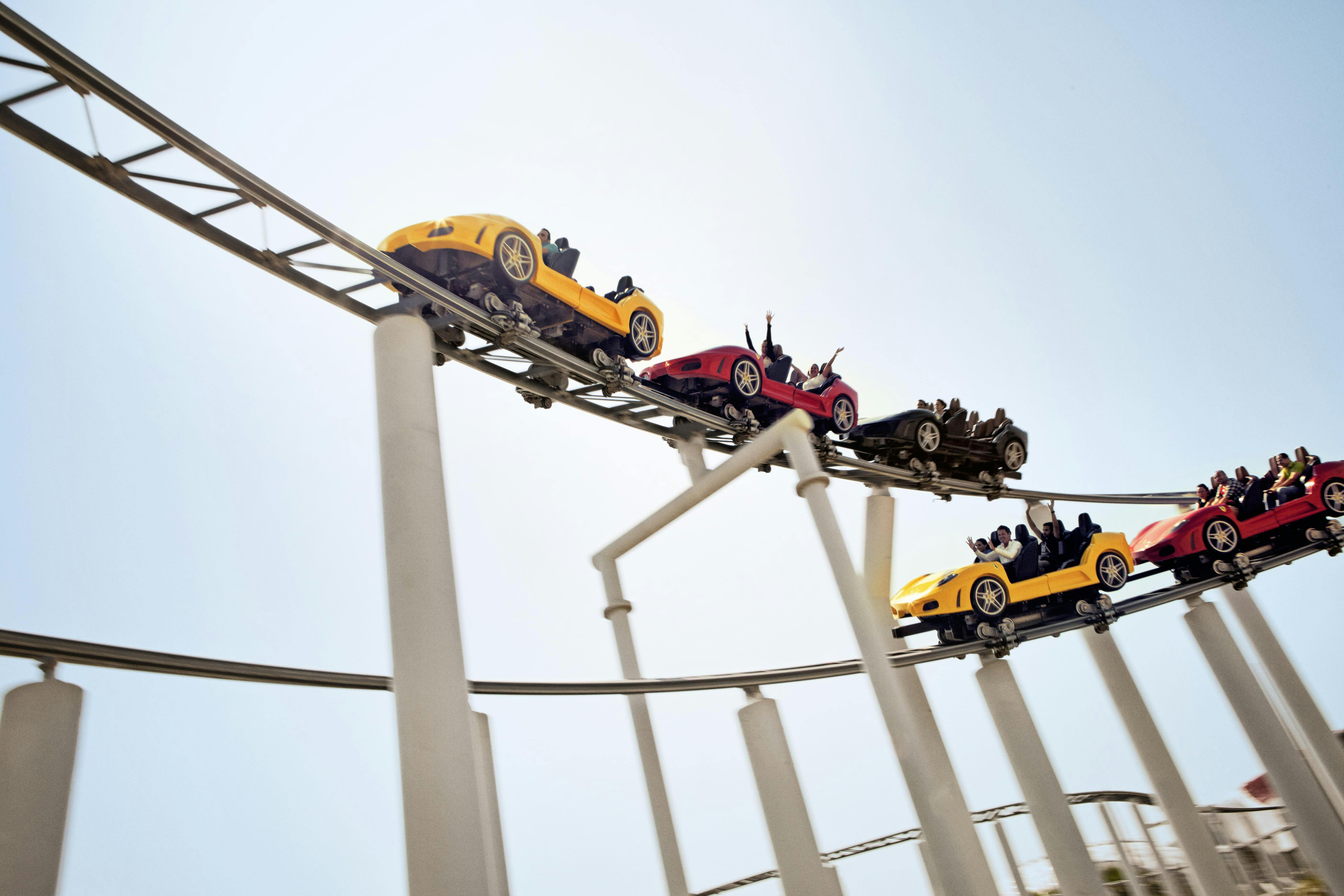 People riding a rollercoaster with car-shaped cars in red, yellow, and black, raising their arms against a clear blue sky.