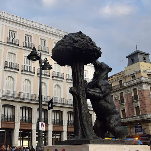 Bronsstaty av en björn som lutar sig mot ett träd framför historiska byggnader med balkonger och människor samlade nedanför.