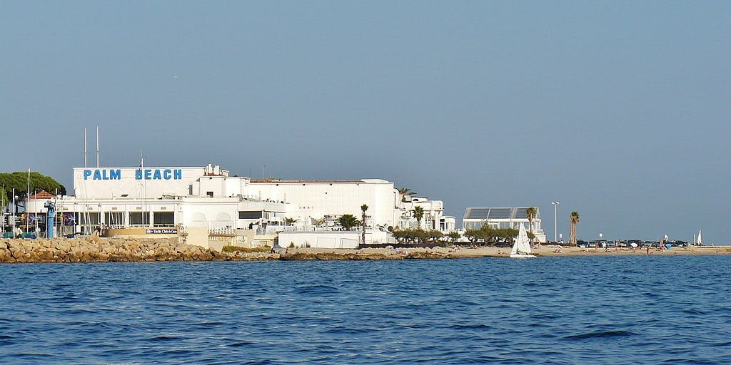 Białe budynki oznaczone „Palm Beach” w pobliżu skalistego wybrzeża i czystej niebieskiej wody, z rzadkimi palmami i kilkoma osobami na piaszczystej plaży.